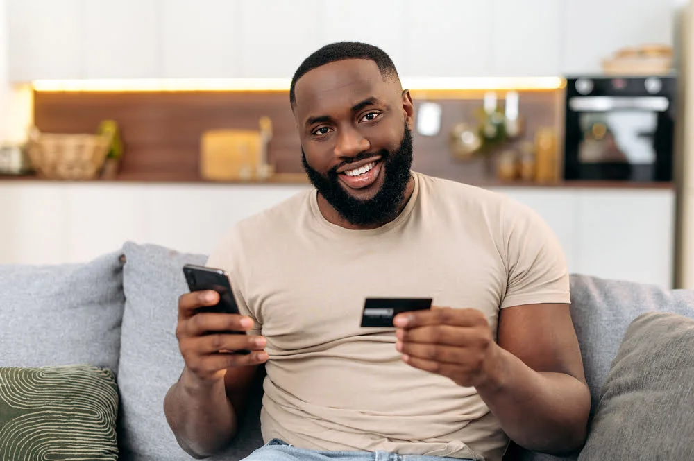 Why Mobile Crypto Casino Games Are Taking the Lead 3 Happy african american attractive man, sits on a sofa in the living room, holding a bank card and a smartphone, making online purchases, making payments, ordering delivery. Online shopping, payment